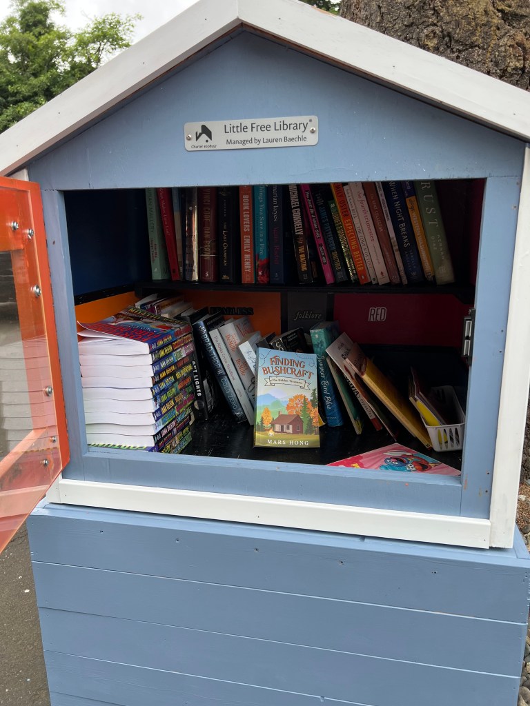 A little free library filled with various books, including the title 'Finding Bushcraft' by Mars Hong, displayed in a colorful wooden structure.