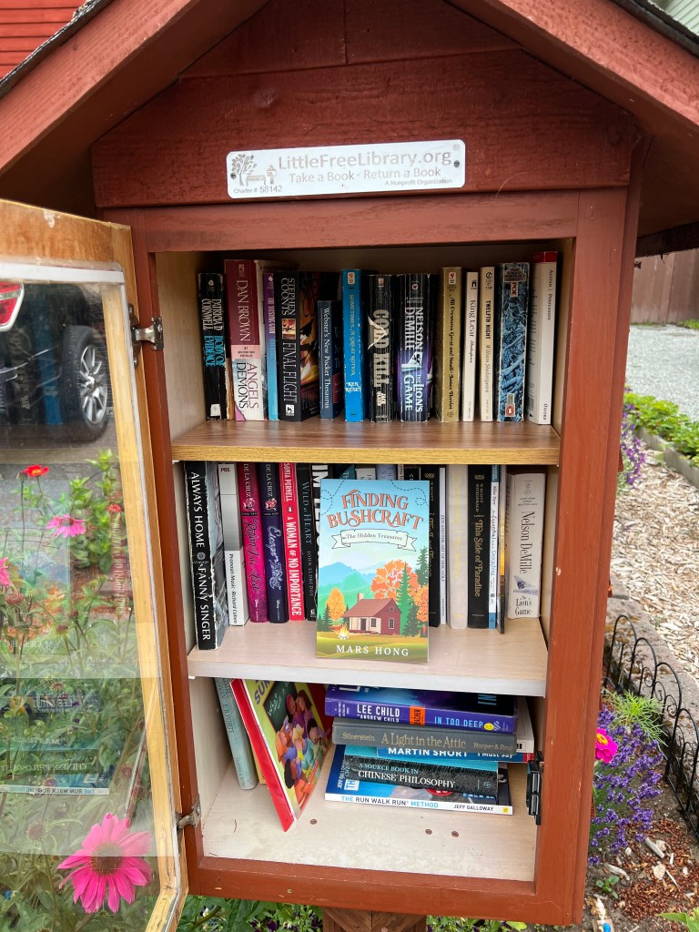 A view of a Little Free Library filled with various books, showcasing titles like 'Finding Bushcraft' by Mars Hong and others, alongside colorful book spines and a wooden structure.