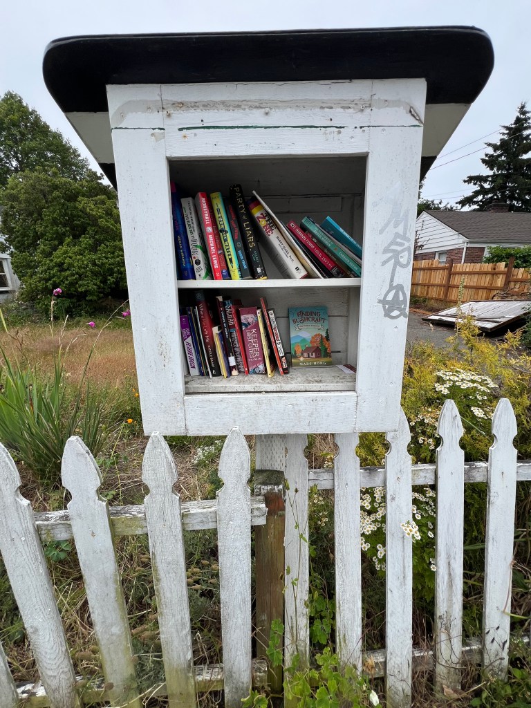 A small, white wooden book exchange box filled with various books, standing beside a white picket fence in a grassy area.