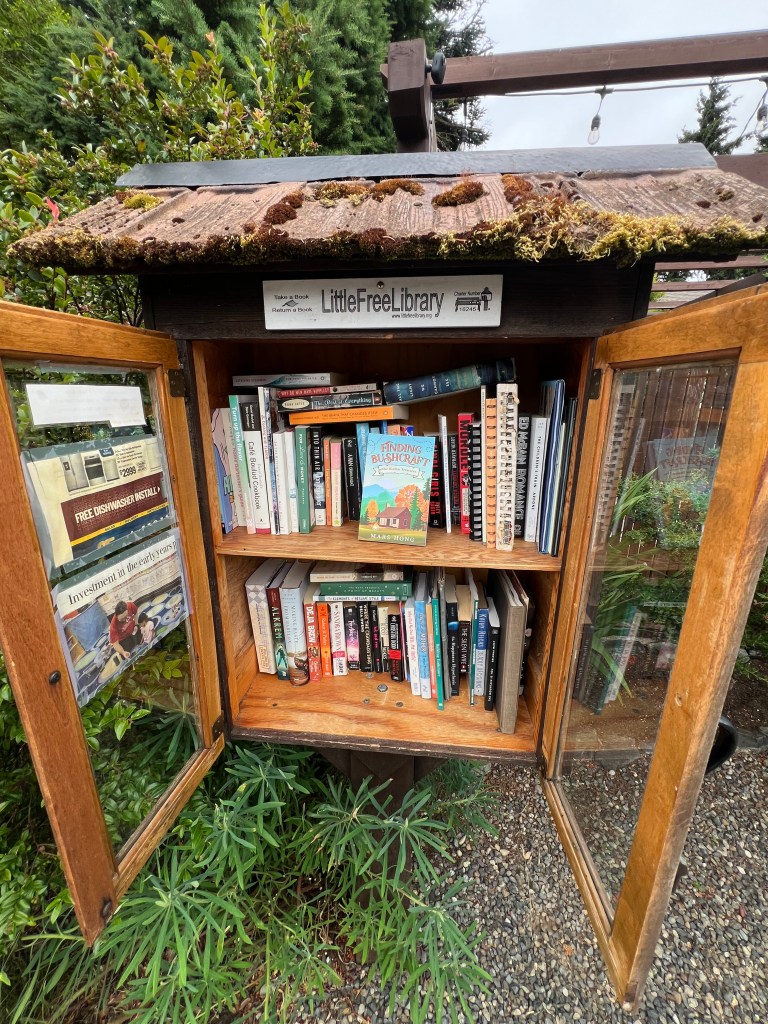 A wooden Little Free Library with double doors, filled with various books, located in a garden setting. The roof is partially covered in moss.