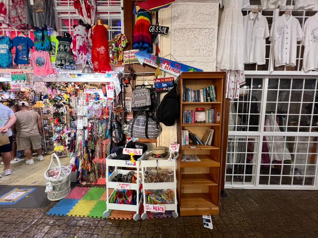 A bustling market scene featuring colorful clothing, souvenirs, and accessories displayed on racks and shelves. A wooden bookshelf filled with books and decorative items is prominently placed, alongside a cart with various trinkets and signs indicating sales. Pedestrians can be seen browsing the vibrant stalls.