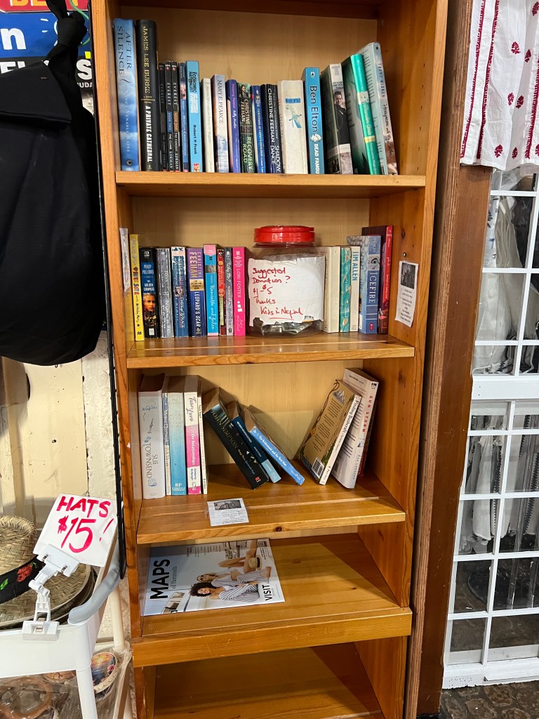 A wooden bookshelf displaying various books across three shelves, with a donation jar and a note suggesting donations. A sign for hats priced at $15 is visible in the foreground.